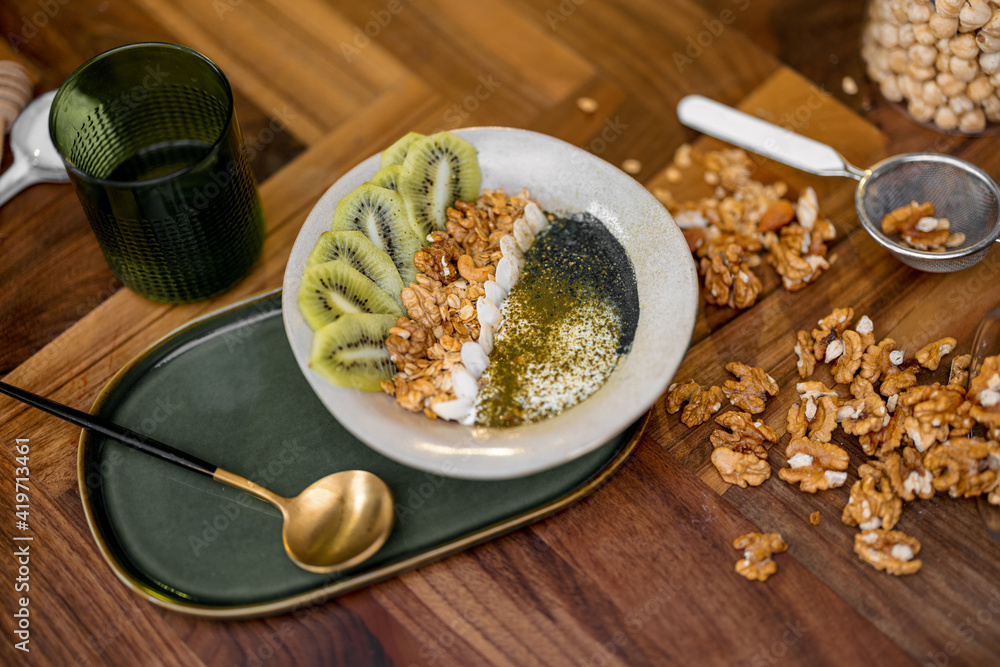 Healthy breakfast with granola bowl with kiwi, spirulina and organic yogurt on wooden table top in kitchen with nuts on background. Healthy breakfast with granola bowl with kiwi, spirulina and organic yogurt on wooden table top in kitchen with nuts on background.