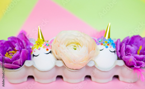 white Easter eggs decorated in the form of unicorns on a colorful background with ranunculus flowers, a minimal creative concept of a happy Easter © klavdiyav