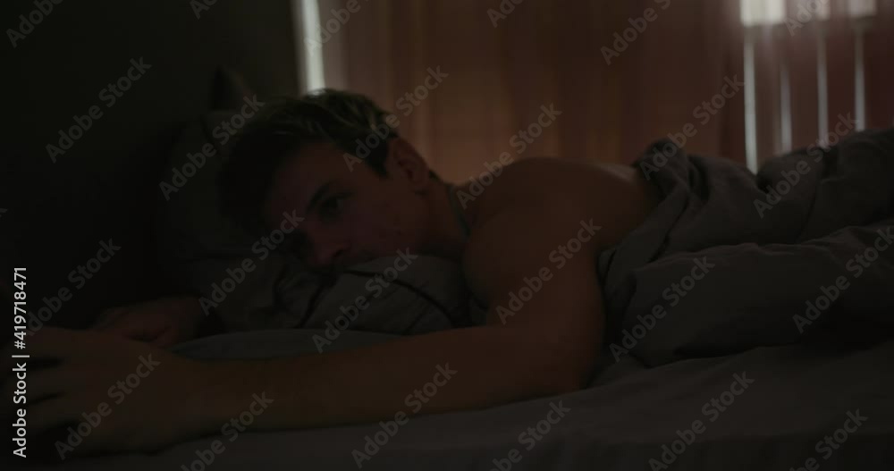 Guy using smartphone on bed in morning. Young man lying on bed and browsing smartphone in morning in dark bedroom at home Guy using smartphone on bed in morning. Young man lying on bed and browsing smartphone in morning in dark bedroom at home