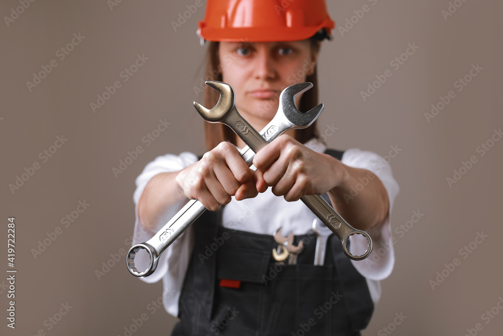 female engineer worker in a uniform and safety helmet with two cross wrenches in her hands and showing in camera. Equality between men and women. Repairwoman posing on a gray background.