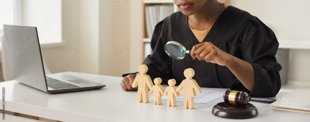 © Studio Romantic - African american judge looking through magnifying glass at wood family figure making decision on children custody. Parental rights deprivation law and guardianship concept. Cropped portrait