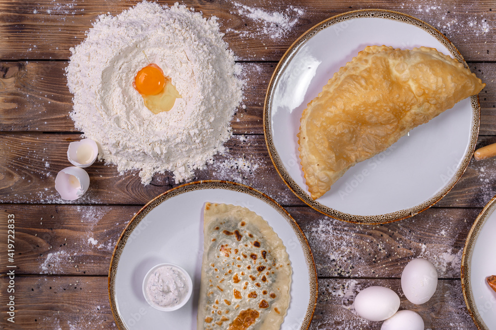 Chebureks on a plate, sauce, flour, chicken eggs on a brown wooden background Chebureks on a plate, sauce, flour, chicken eggs on a brown wooden background