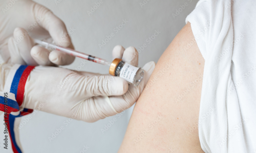 Open shoulder of the man before vaccination. Medical assistant preparing the syringe and Russian flag on her hand in the background. Open shoulder of the man before vaccination. Medical assistant preparing the syringe and Russian flag on her hand in the background.