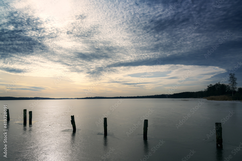 Row of dark wooden piles on a frozen lake under a sky with interesting clouds, in late winter the earth stands still while there is already movement in the atmosphere preparing for spring, copy space,