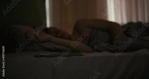 Male turning off alarm clock on smartphone. Young guy waking up and turning off alarm clock on mobile phone while lying on bed early in morning in dark bedroom