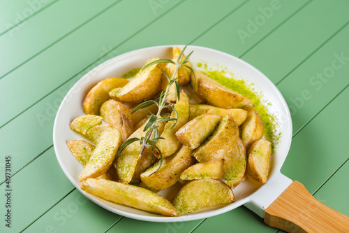Baked potatoes with green sauce and rosemary on a white plate in the form of a frying pan. Fried potatoes with pesto sauce on a green table.