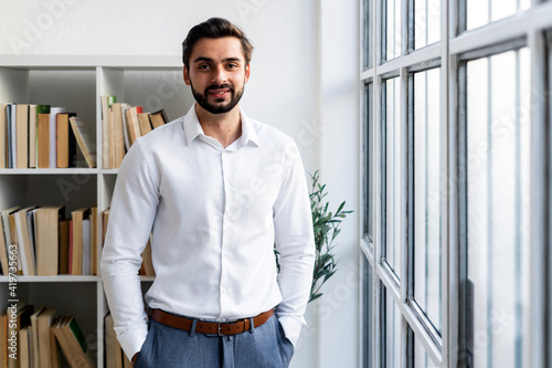 Smiling businessman with hands in pockets standing against bookshelf in office