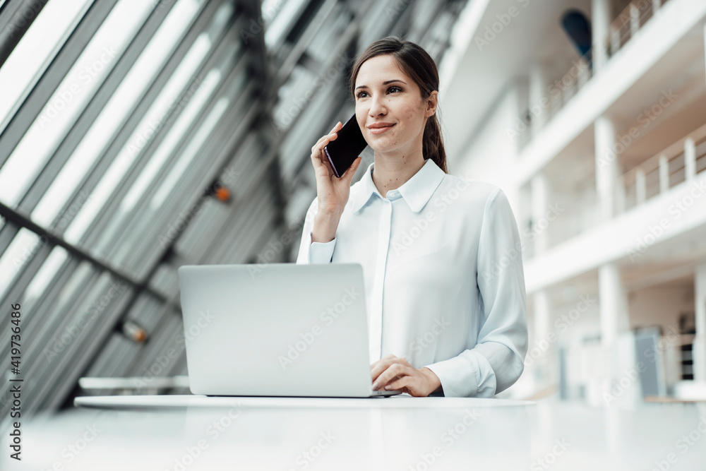 Smiling female entrepreneur talking on smart phone while working on laptop in corridor Smiling female entrepreneur talking on smart phone while working on laptop in corridor