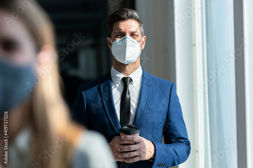 Businessman wearing protective face mask holding coffee while standing in office photo