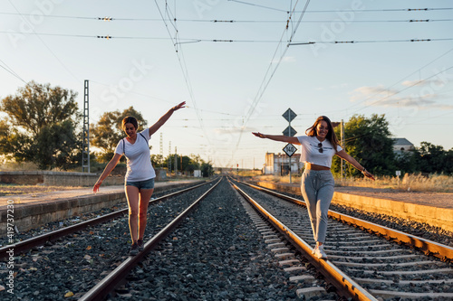 Carefree female friends with arms outstretched walking on railroad track against sky at sunset