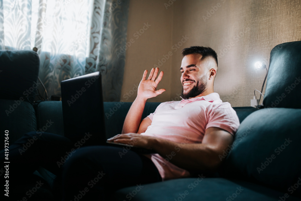 Young businessman waving hand on video call through laptop while sitting on couch in living room Young businessman waving hand on video call through laptop while sitting on couch in living room