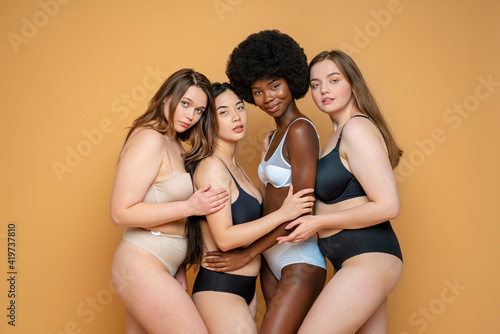 Multi ethnic female model friends in lingerie embracing each other against yellow background