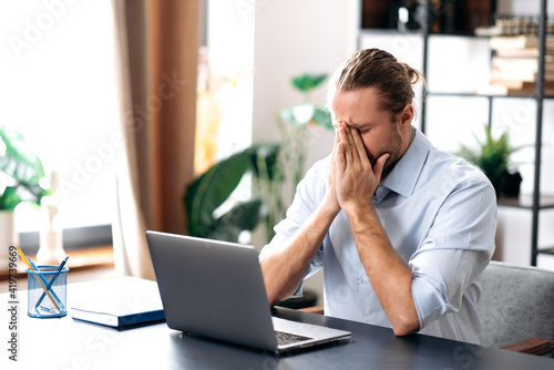 Caucasian unhealthy man feeling bad, sick with running nose, need a rest.Male manager or freelancer sit at the desk at home office, working distantly, using paper tissue, sneezing