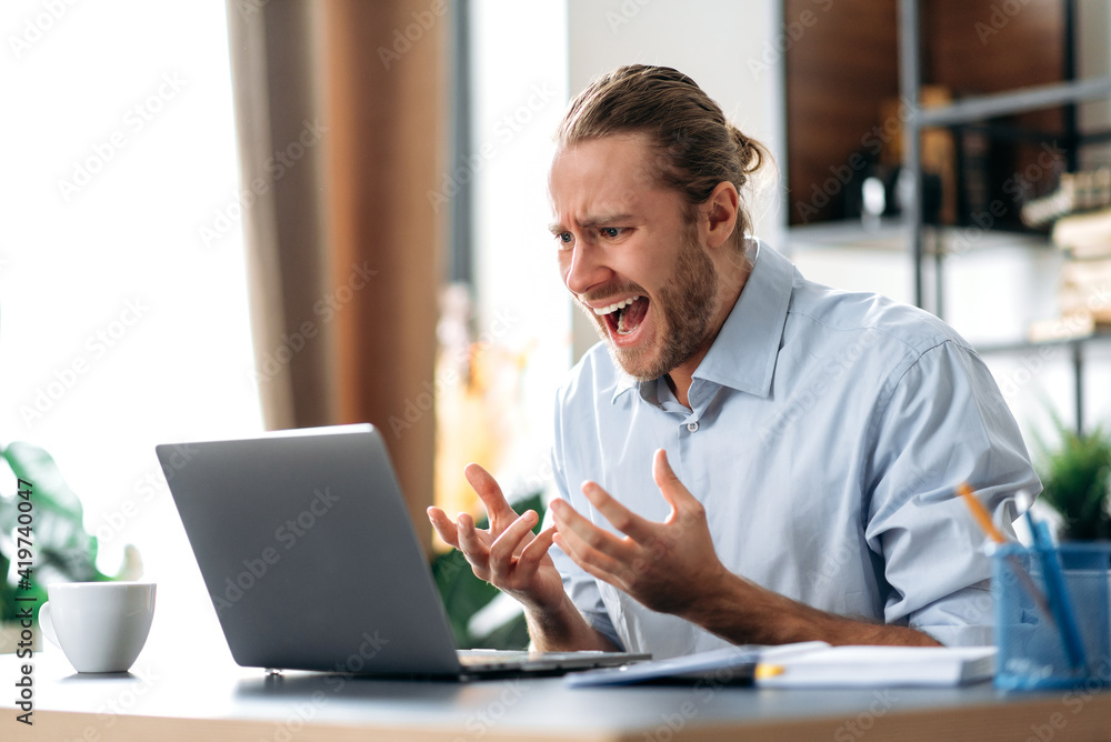 Shocked annoyed caucasian busy guy looking at a laptop in confusion, received unexpected news, failed a deal, shouts, gesturing with hands, sitting at the work desk at home office Shocked annoyed caucasian busy guy looking at a laptop in confusion, received unexpected news, failed a deal, shouts, gesturing with hands, sitting at the work desk at home office