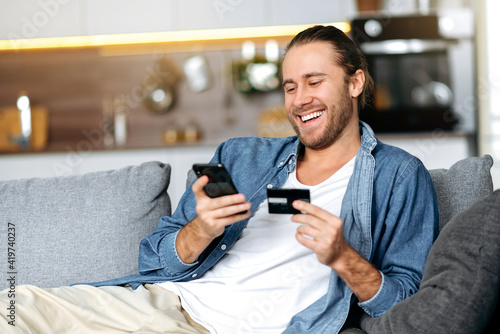 A happy smiling caucasian guy, in casual stylish clothes, sits on the couch at home, holds a phone and a banking card in his hands, makes online payment for purchases, enters a card number