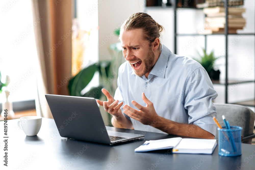 Annoyed caucasian busy business guy looking at a laptop in confusion, received unexpected news, failed a deal, shouts, gesturing with hands, sitting at the work desk at home office Annoyed caucasian busy business guy looking at a laptop in confusion, received unexpected news, failed a deal, shouts, gesturing with hands, sitting at the work desk at home office