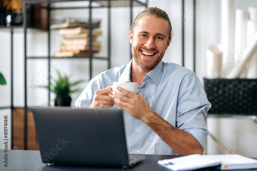Happy friendly confident caucasian guy, employee or freelancer, holding a cup of coffee while sitting at his desk at home or office, looking at the camera with a friendly smile © Kateryna