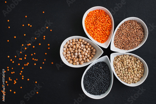 Different types of lentils in ceramic bowls (red turkish lentils, canadian lentils, black lentils - beluga, buckwheat, chickpeas) on black slate table.. For culinary, shop, restaurant. Top view .
