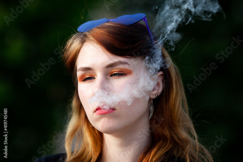 Vape teenager. Young pretty white caucasian girl with glasses on her head an electronic cigarette on the street in the summer. Deadly bad habit. © aleksandr_yu