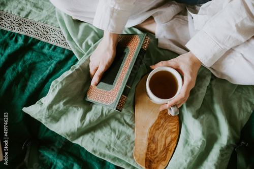Women's hands hold a hot cup of tea on the bed, linen bedding, country life, reading in bed, relaxation, tranquility, digital detox. High quality photo