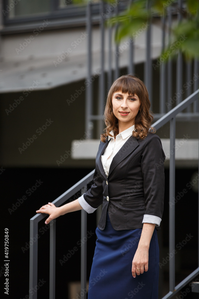 Fototapeta premium Young woman in classic business suit on background of a City park. Business style girl in Summer City on background of new park near a building. Street style Modern strict urban girl.
