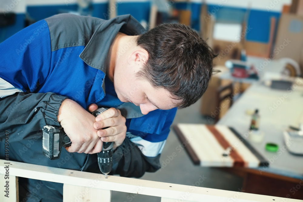 A young worker in overalls with a screwdriver collects furniture in a carpenter's shop. A white man screws a self-tapping screw into wooden planks. Background for making furniture. A young worker in overalls with a screwdriver collects furniture in a carpenter's shop. A white man screws a self-tapping screw into wooden planks. Background for making furniture.