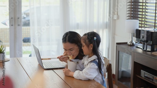 Asian family young mother working on laptop computer and her little daughter bring instant noodles to mom at home, woman eats noodles while doing work from home