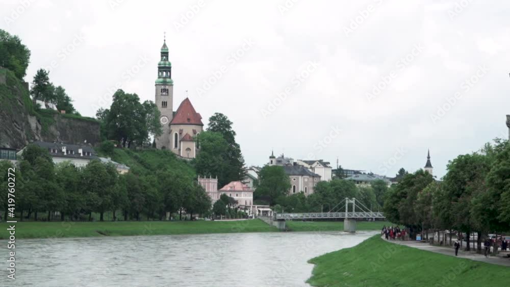 View Of Pfarre Mulln In Salzburg, Austria With Mullner Steg Bridge Across Salzach River. wide shot View Of Pfarre Mulln In Salzburg, Austria With Mullner Steg Bridge Across Salzach River. wide shot