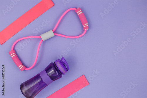Fitness clothes and accessories for woman on violet background. Sports fashion with t-shirt, elastic bands, headphones, phone, bottle. Healthy, active lifestyle concept