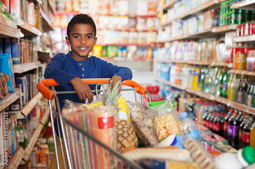 Positive preteen African boy carrying full grocery cart after shopping in food store