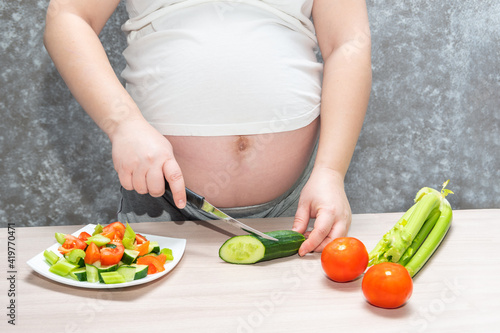 Pregnant woman cutting cucumber for fresh green salad, female prepares tasty organic dinner at home, healthy nutrition for future mother. Concept of healthy lifestyle and nutrition during pregnancy