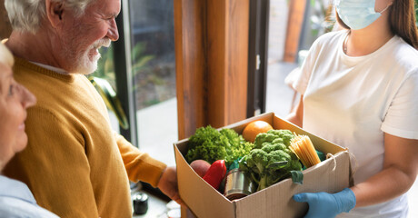 Food delivery or donation concept. Young woman in gloves and protective face mask delivers a box of food elderly couple in home