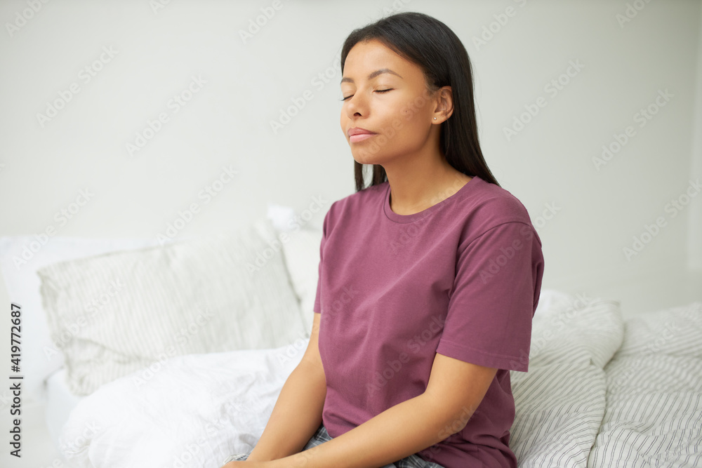 Portrait of adorable beautiful young dark haired black woman sitting on bed wearing pajamas keeping eyes closed having calm peaceful facial expression practicing mindfulness meditation in the morning Portrait of adorable beautiful young dark haired black woman sitting on bed wearing pajamas keeping eyes closed having calm peaceful facial expression practicing mindfulness meditation in the morning
