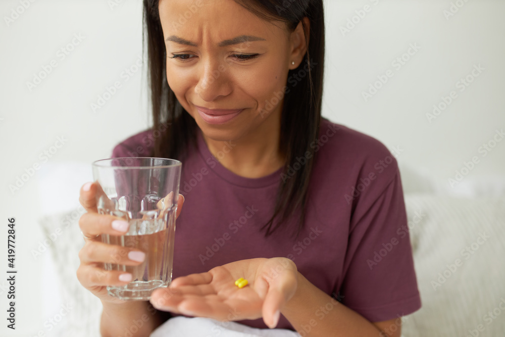 Attractive emotional young Afro American female in pajamas posing at home, holding glass of pure water and pills, taking daily dosage of vitamins or food supplements for beauty skin and hair Attractive emotional young Afro American female in pajamas posing at home, holding glass of pure water and pills, taking daily dosage of vitamins or food supplements for beauty skin and hair