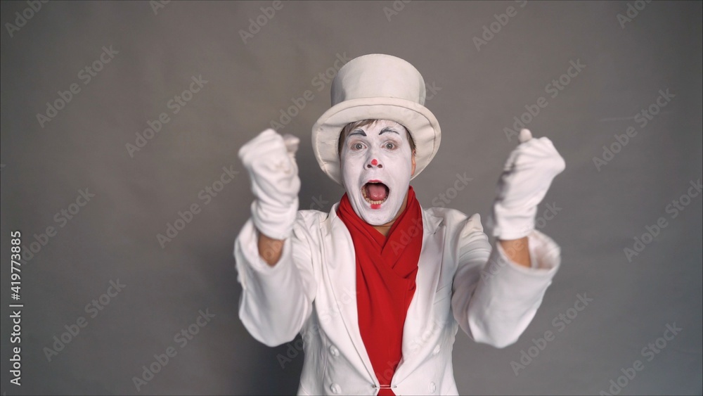 Joyful pantomime. Mime in a white suit and hat is very pleased. The  surprised face of a clown-mime in a white hat. The clown rejoices on a gray  background. Stock Photo