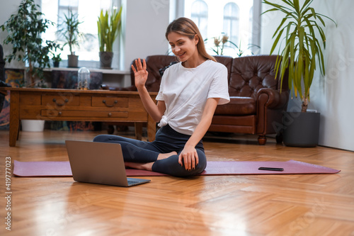 Female sportive blogger smiles and greets her followers on her laptop sitting on mat in modern living room.