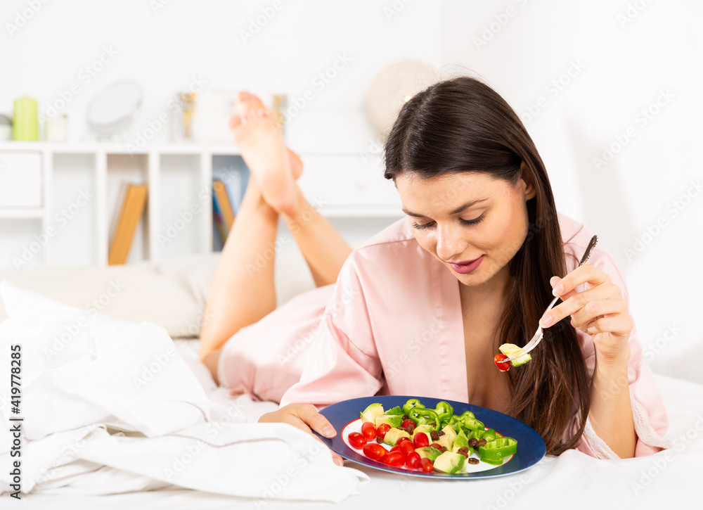 Young pretty woman in bathrobe eating vegetable salad from plato in bed at home