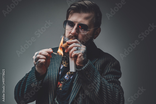 Portrait of a serious and handsome man with stylish coiffure posing in gray background. Tattooed man smoking cigar.