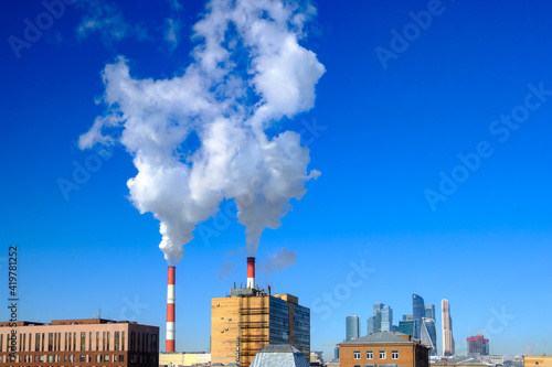White puffs of smoke rise into the blue cloudless sky from industrial chimneys above the rooftops of houses in the city. Sunny frosty day in winter.