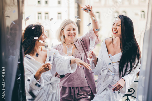 Pretty young women friends in bathrobes and pajama clink elegant glasses with delicious champagne at bachelorette party on hotel room terrace