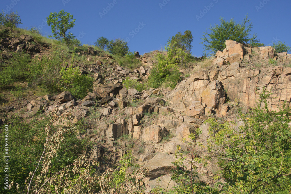 A rocky slope of an abandoned granite quarry in the Nikolaev region of Ukraine on a hot summer day. Sharp stones overgrown with sparse vegetation. Blue cloudless sky over the cliffs.