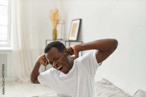 Portrait of sleepy young black male in nightwear sitting on bed in cozy room opening mouth, closing eyes and stretching body right after awakening, yawning, feeling lazy to get up so early
