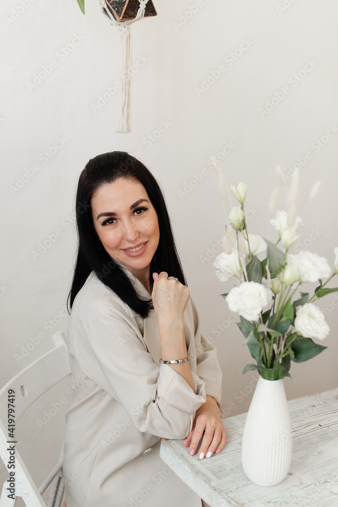 brunette girl sitting at a white table with flowers in a vase brunette girl sitting at a white table with flowers in a vase