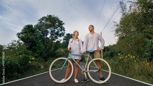 Fototapeta Naklejka Na Ścianę i Meble -  Biking road trip. Love couple on blue bike in white clothes on forest road. Just married woman and man kiss, hugs, stand on bicycle. Wedding, honeymoon. Cycling Cycle Fix. Asia Thailand ride tourism.