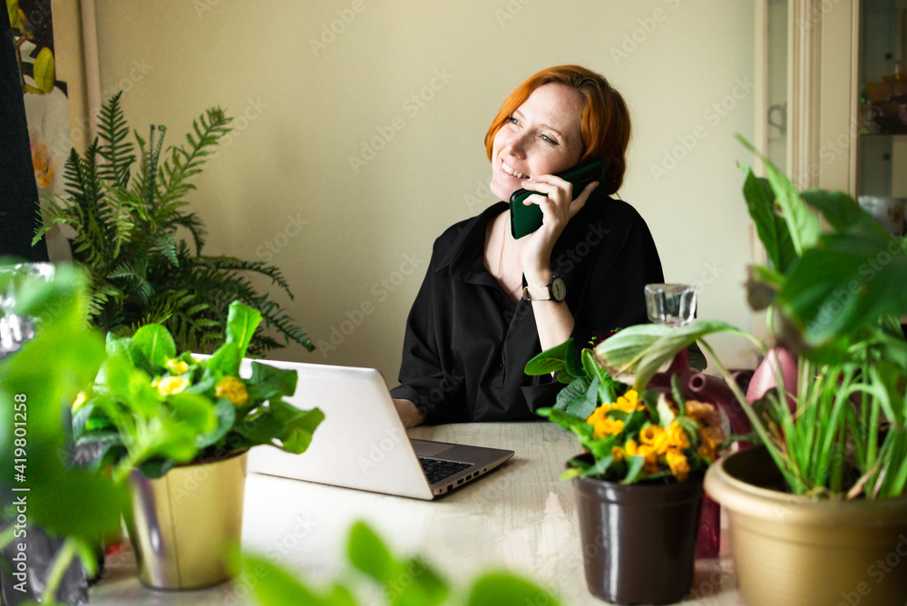 Smiling red-haired woman working on laptop and talking on the phone among home garden and houseplants. Concept of remote work from home, quarantine, working woman freelancer. Home Office. Smiling red-haired woman working on laptop and talking on the phone among home garden and houseplants. Concept of remote work from home, quarantine, working woman freelancer. Home Office.