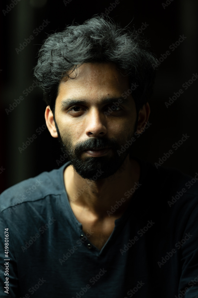 Young handsome Indian Asian male boy with black dense hair and beard looking at the camera to pose. Dark and moody theme with lights and shadows on face isolated on black background