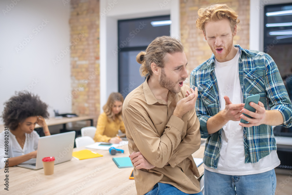 Fototapeta premium Two male colleagues discussing something while looking at the phone abd looking enjoyed