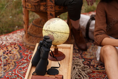 Eine Frau im Safari look sitzt draussen auf dem Tepich und  liest ein altes Buch, auf der Holzkiste steht ein Globus und ein fernglas.Sie ist auf der Reise, liebt Abendteuer