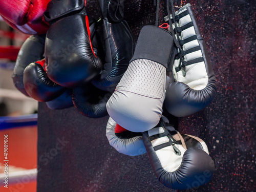 A bunch of boxing gloves hanging on a gym wall with boxing ring in the background. Black and white boxing gloves for training and fighting. Sport equipment for kickboxing and martial arts