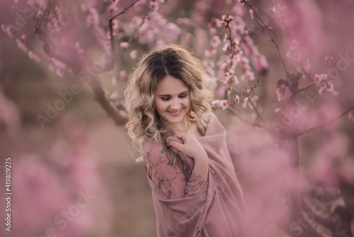 Gentle portrait Young curly blonde woman in brown pleated skirt pink blouse covered shoulders with stole stands in blooming peach gardens at sunset. Girl enjoying walk outside the city, natural beauty